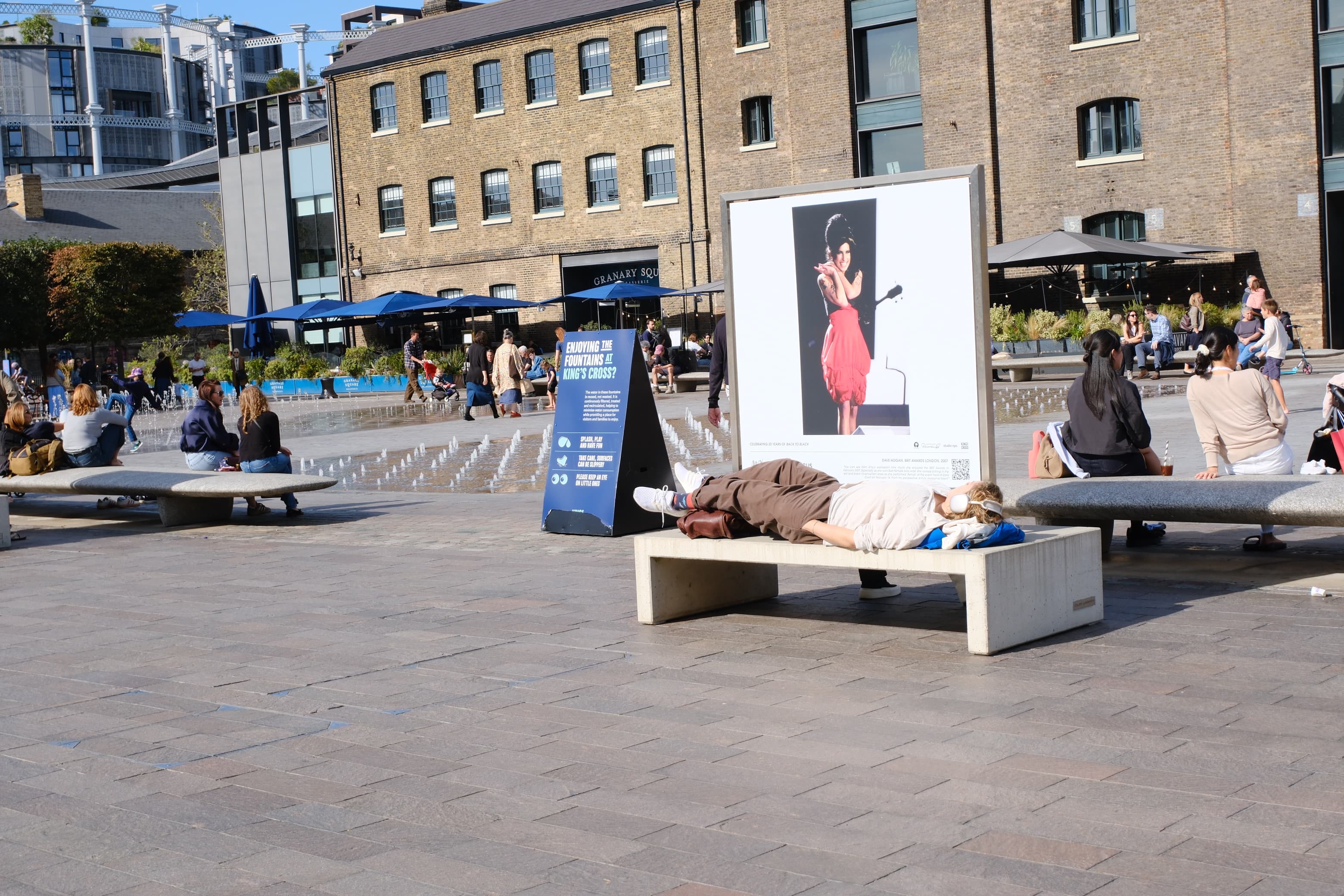 Someone napping on a concrete bench in headphones beside a large exhibition photo, fountains behind