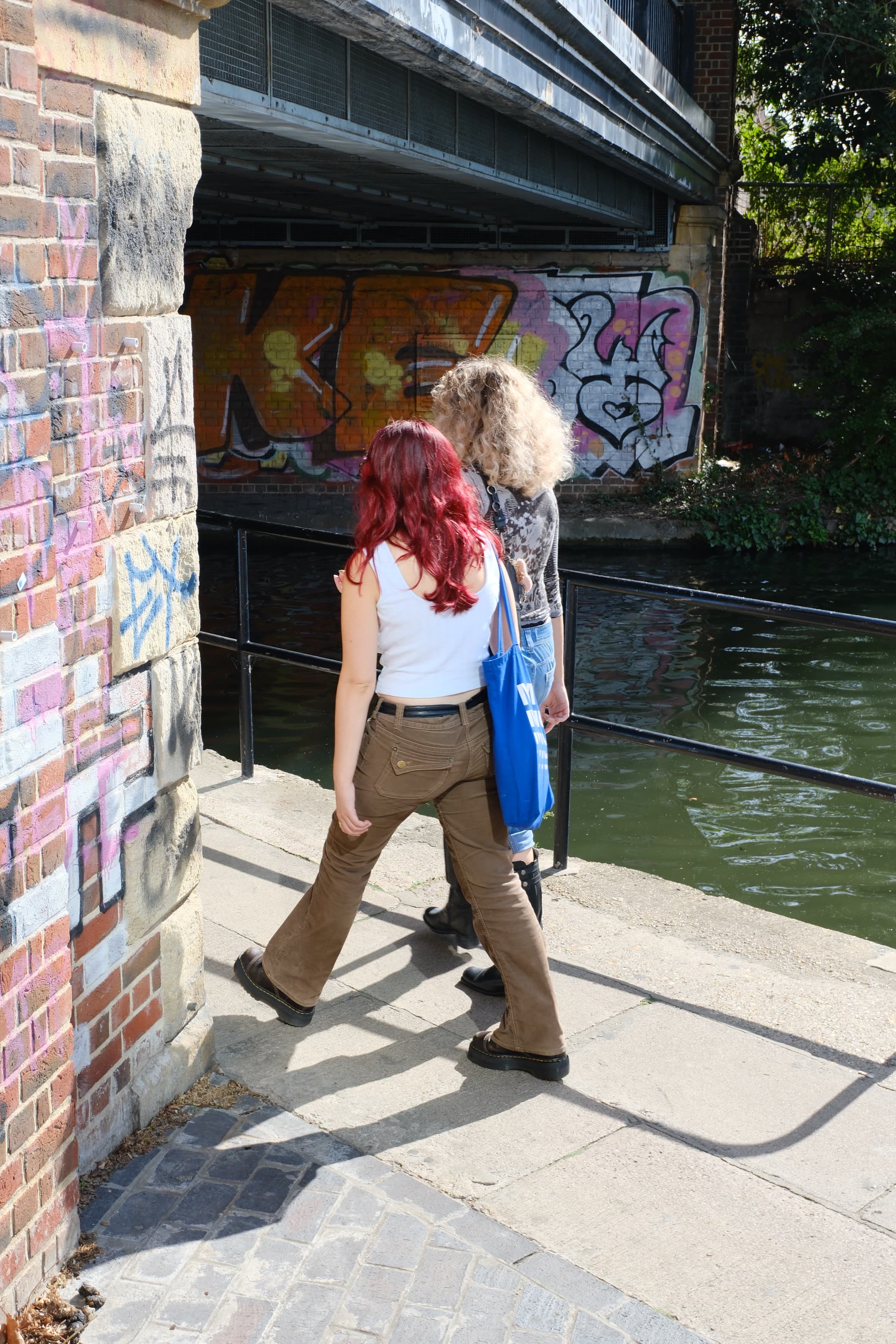 Two friends walking along the canal towpath under a graffitied bridge, one with red hair and a blue tote bag