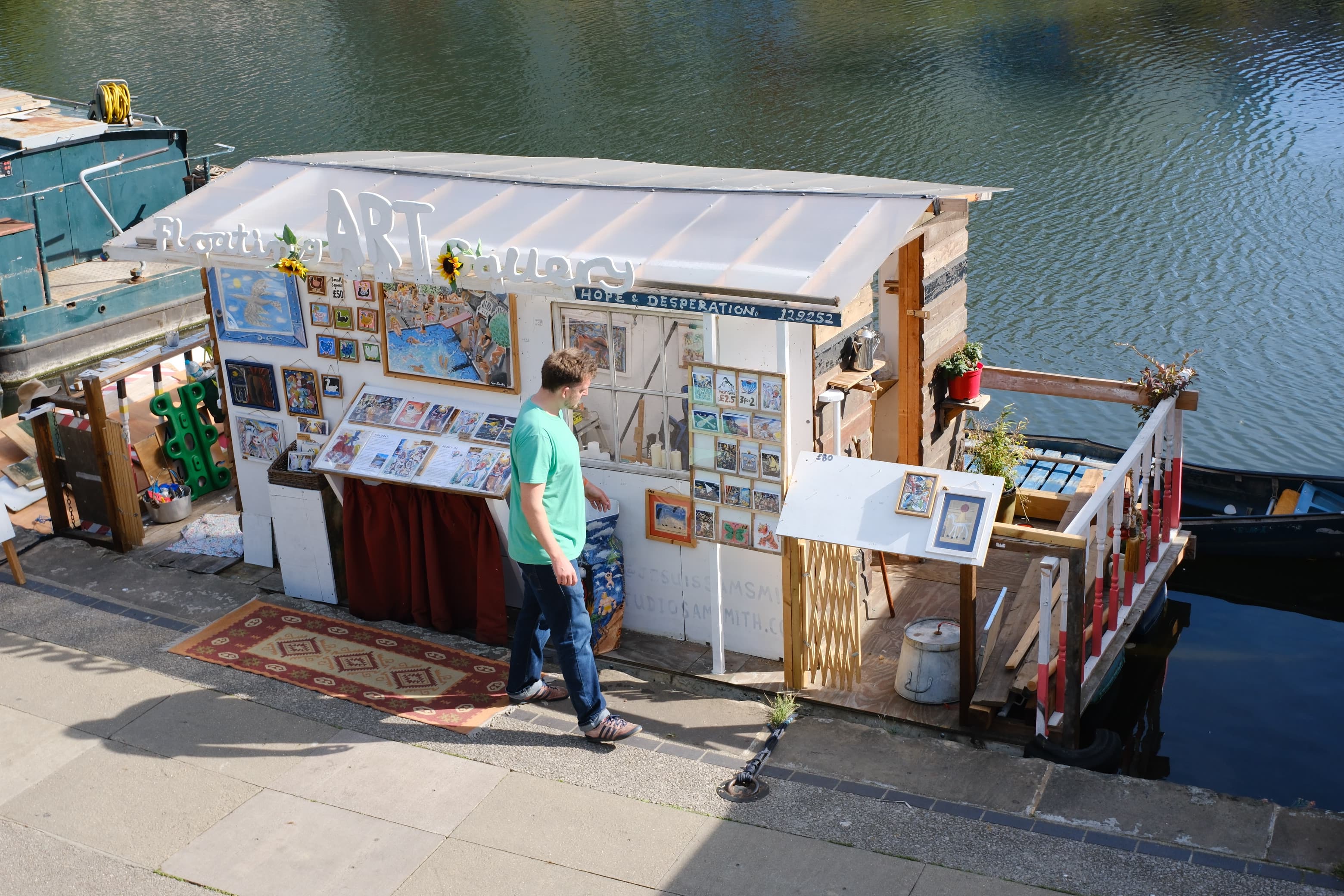 A man in a green T-shirt browsing paintings outside a floating art gallery moored on the canal