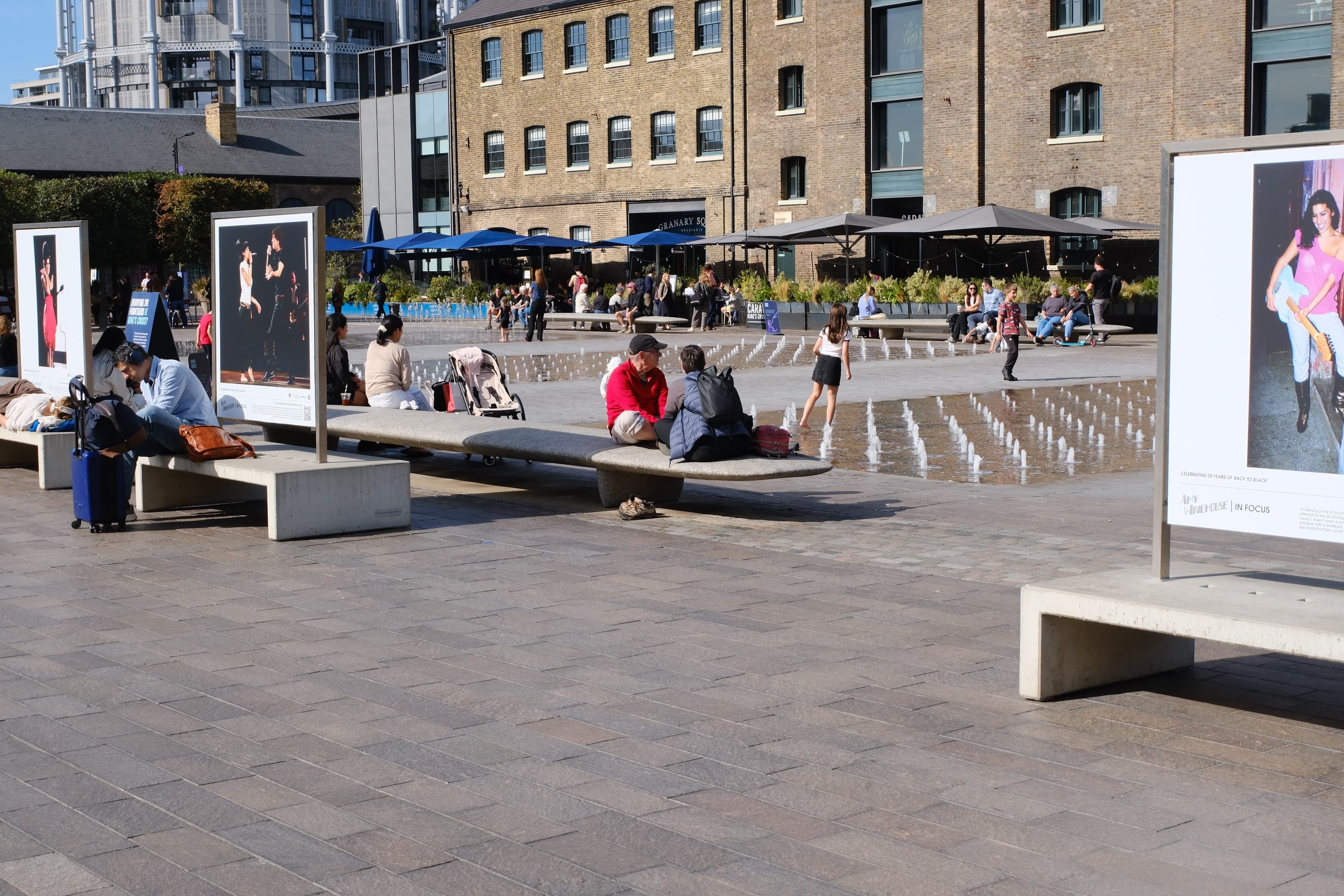 People sitting on stone benches beside the fountains in Granary Square, a photo exhibition on display